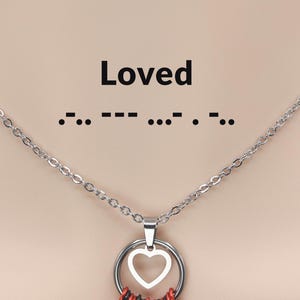 Puede incluir: Un collar de plata con un colgante en forma de corazón. El colgante está rodeado por un círculo de anillos rojos y negros. El collar tiene la palabra "Loved" y la palabra "Loved" escrita en código Morse.