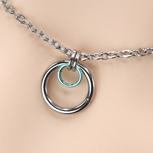 Puede incluir: Un collar de cadena de plata con un colgante de anillo grande de plata. El anillo tiene un anillo verde más pequeño en su interior.