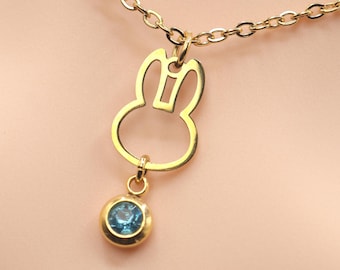 May include: A gold-colored necklace featuring a bunny pendant with a blue gemstone. The bunny pendant is a cutout design, and the gemstone is round. The necklace is on a light pink background.