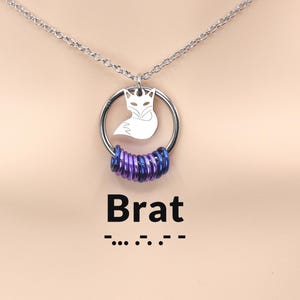 May include: A silver-toned chain necklace featuring a circular pendant with a fox silhouette cutout. The bottom of the ring is adorned with a series of small, iridescent purple and blue metallic jump rings. The text Brat is printed below the pendant.
