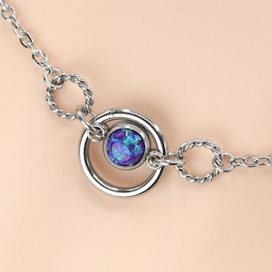 Puede incluir: Un collar plateado con un colgante central que presenta una piedra preciosa azul y púrpura brillante. El colgante está enmarcado por un anillo de metal pulido, conectado a una fina cadena con detalles retorcidos. La joya se encuentra sobre un fondo neutro.