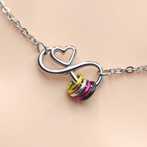 May include: A silver-toned chain necklace featuring a central infinity symbol charm with an integrated heart shape. Three small, colorful rings in yellow, magenta, and black are suspended from the bottom loop of the infinity symbol charm.