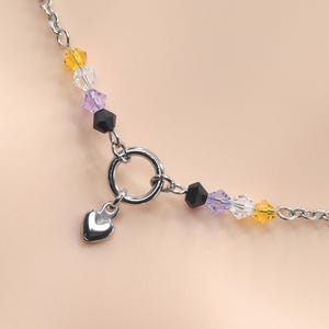 May include: A silver-toned chain necklace featuring a central circular clasp with a small, smooth heart-shaped charm. The chain is accented on both sides of the clasp with a sequence of faceted beads in yellow, clear, light purple, and black colors.