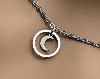 Könnte beinhalten: Silberfarbene Halskette mit einem Mondsichel-Anhänger in einem kreisförmigen Rahmen. Die Kette ist filigran und der Anhänger ist mittig platziert, wodurch ein minimalistisches und elegantes Design entsteht. Die Halskette wird vor einem neutralen Hintergrund gezeigt.