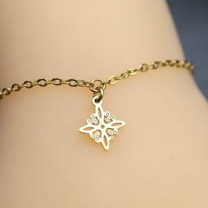 Könnte beinhalten: Ein goldfarbenes Metallkettenarmband mit einem einzelnen, zentralen Anhänger. Der Anhänger hat eine symmetrische, vierzackige Form mit komplizierten, durchbrochenen Schnörkelmustern und Wirbeldetails, die an einer feinen Gliederkette hängen.