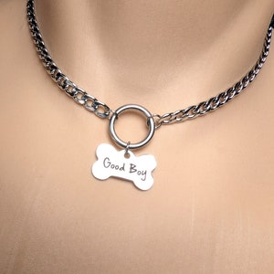 Puede incluir: Un collar gargantilla de cadena plateada con un anillo grande de plata y un dije de hueso plateado que dice "Good Boy".