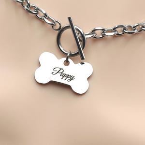 May include: A silver-colored dog collar with a bone-shaped tag. The tag is engraved with the word "Puppy" in a cursive font. The collar has a toggle clasp and a chain link design.