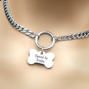 Puede incluir: Un collar gargantilla de cadena plateada con un anillo grande de plata y un dije de hueso plateado que dice "Owned by Daddy".