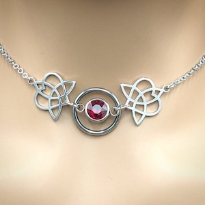 Könnte beinhalten: Eine silberne Halskette mit einem keltischen Knotendesign auf jeder Seite eines runden Anhängers mit einem roten Edelstein in der Mitte.