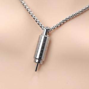 May include: A silver-toned metal pendant shaped like a small, segmented cylinder with a thin pin extending from the bottom. The pendant hangs from a matching silver-toned wheat chain necklace, featuring a polished finish and a sleek, industrial design.