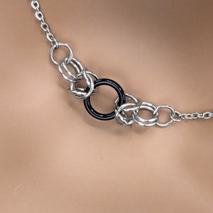 Pode incluir: Um colar de corrente de prata com um pingente de anel preto. A corrente é feita de anéis entrelaçados.