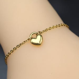 Könnte beinhalten: Ein goldfarbenes Kettenarmband mit einem einzelnen, glatten, polierten herzförmigen Anhänger, der in der Mitte hängt. Die feinen Metallglieder bilden eine durchgehende Schlaufe und zeigen ein schlichtes Design mit einer reflektierenden Oberfläche.