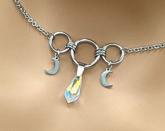 Puede incluir: Un collar de cadena de plata con tres anillos conectados y dos dijes de luna creciente. Un cristal en forma de lágrima cuelga del anillo central.