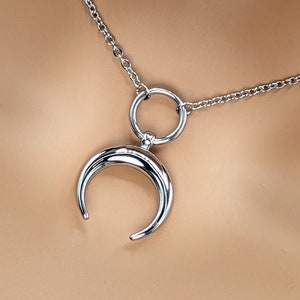 Puede incluir: Un collar de plata con un colgante de luna creciente y un dije circular. La cadena es de estilo delicado y enlazado.