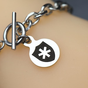 May include: A silver-toned metal chain bracelet featuring a toggle clasp and a circular charm. The charm displays a black shield-shaped emblem with a white asterisk symbol centered within it. The bracelet is constructed from polished metal links.
