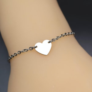 Könnte beinhalten: Ein Armband aus einer dunkelmetallischen Kette mit einem einzelnen, glatten, silberfarbenen Herzanhänger, der mittig an den Gliedern befestigt ist. Das Armband besteht aus einer einfachen Rundgliederkette, die an beiden Seiten des flachen Herzanhängers ansetzt.