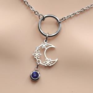 May include: A silver necklace with a crescent moon pendant decorated with floral cutouts and a dangling blue gemstone. The necklace has a fine chain and a circular ring element at the top.