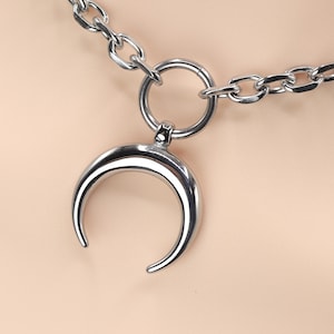 Könnte beinhalten: Silberfarbene Halskette mit einem Halbmond-Anhänger. Die Kette hat ein Kettenglied-Design mit einem großen kreisförmigen Ring, der die Kette mit dem Mond-Anhänger verbindet. Der Mond-Anhänger ist glatt und gebogen.