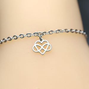 Könnte beinhalten: Ein silberfarbenes Metallkettenarmband mit einem einzelnen Anhänger in Form eines Herzens, das mit einem Unendlichkeitssymbol kombiniert ist. Die feine Gliederkette ist poliert und glänzend, wobei der Anhänger mittig an einem Ring hängt.