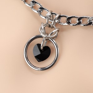Può includere: Collana a catena color argento con un ciondolo a cuore nero. Il ciondolo è un cuore nero sfaccettato all'interno di un anello d'argento. La catena è a maglia grossa, su sfondo neutro.