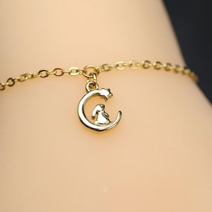 Könnte beinhalten: Ein goldfarbenes Kettenarmband mit einem kleinen Anhänger in Form einer Mondsichel, der oben einen winzigen Stern und eine kleine Hasenfigur in der Krümmung des Mondes zeigt. Das Metall hat durchgehend eine polierte, glänzende Oberfläche.