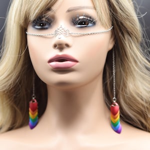 Puede incluir: Un aro de nariz de cadena plateada con un diseño decorativo. La cadena está unida a pendientes de colores arcoíris con un diseño en forma de corazón.