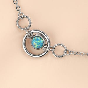 Könnte beinhalten: Eine silberfarbene Halskette mit einem zentralen Anhänger. Der Anhänger hat einen blau-grünen, schillernden Stein, der in einem silbernen Ring gefasst ist. Die Kette hat verdrehte Metallglieder. Die Halskette ist zum Tragen um den Hals konzipiert.