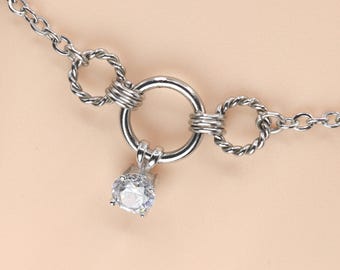 Puede incluir: Collar plateado con un diseño de anillo central y dos anillos trenzados más pequeños a cada lado. Un colgante de gema transparente y redonda cuelga del anillo central. La cadena es delicada.