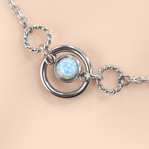 May include: A silver-toned necklace featuring a central circular pendant with a light blue opal-like stone. The necklace has a delicate chain with twisted ring accents, creating a minimalist and elegant design. Jewelry.