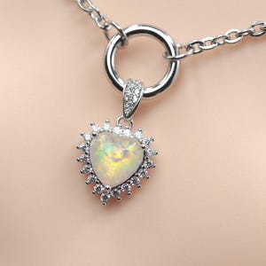 May include: A silver-toned necklace featuring a heart-shaped opal pendant surrounded by a halo of small clear stones. The pendant is attached to a circular silver-toned ring, which connects to a delicate silver-toned chain link necklace.