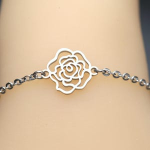Könnte beinhalten: Ein silberfarbenes Metallarmband mit einem mittigen, durchbrochenen Rosenanhänger. Der Anhänger ist beidseitig mit einer feinen Gliederkette aus runden Elementen verbunden, die ein durchgehendes Band bildet und eine polierte Oberfläche aufweist.