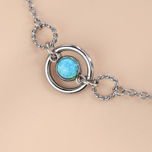 May include: A silver-toned necklace featuring a central pendant with a round, light blue opal stone. The pendant is encircled by a polished silver ring, connected to a delicate chain with twisted and circular links. The opal has a shimmering, iridescent quality.