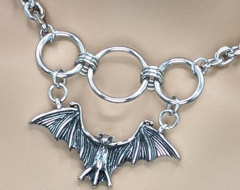 May include: A silver chain necklace with a bat pendant and three interconnected silver rings.