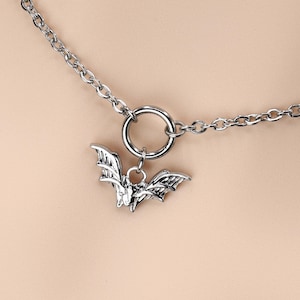 Może przedstawiać: Srebrny naszyjnik typu choker z wisiorkiem w kształcie nietoperza. Naszyjnik ma łańcuszek z dużym pierścieniem i szczegółowym wisiorkiem w kształcie nietoperza. Wisiorek nietoperza jest zawieszony na pierścieniu.