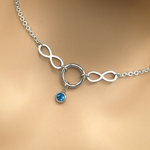 Könnte beinhalten: Eine silberfarbene Halskette mit Unendlichkeitszeichen und einem zentralen Ring. Ein kleiner, runder, blauer Edelsteinanhänger hängt an dem Ring. Die Halskette ist zum Tragen um den Hals konzipiert, wahrscheinlich aus Edelstahl.