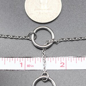 Może przedstawiać: Srebrny naszyjnik z łańcuszkiem i dwoma srebrnymi pierścieniami. Łańcuszek ma około 2,5 cm długości, a pierścienie mają średnicę około 1,25 cm. Naszyjnik jest mierzony za pomocą linijki.