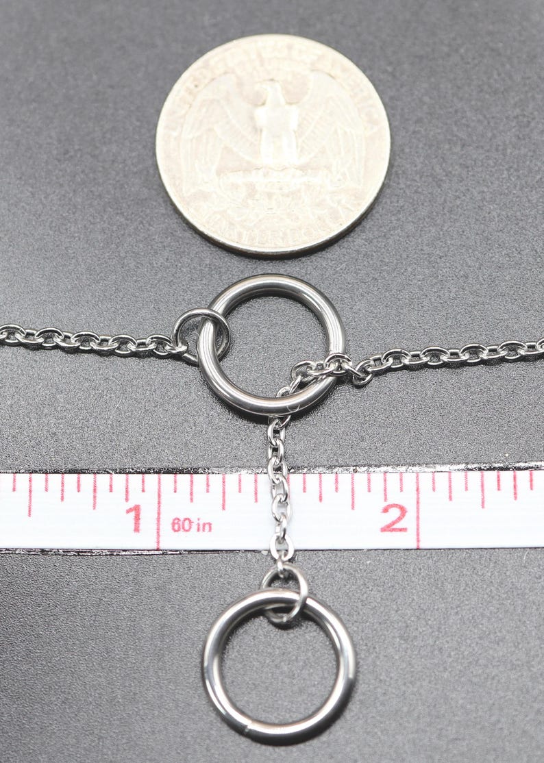 Może przedstawiać: Srebrny naszyjnik z łańcuszkiem i dwoma srebrnymi pierścieniami. Łańcuszek ma około 2,5 cm długości, a pierścienie mają średnicę około 1,25 cm. Naszyjnik jest mierzony za pomocą linijki.