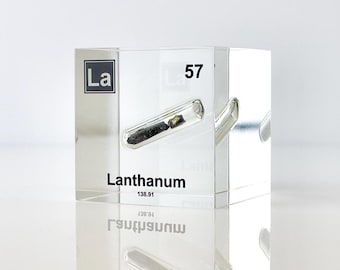 May include: A clear acrylic cube containing a small sample of lanthanum metal. The cube has the element's symbol "La" and atomic number "57" printed on it. The text "Lanthanum 138.91" is also printed on the cube.