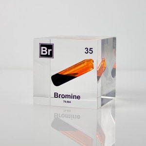 Może przedstawiać: Przezroczysta akrylowa kostka z pierwiastkiem bromu. Kostka wyświetla symbol "Br", liczbę atomową 35 i nazwę "Brom". Wewnątrz kostki znajduje się próbka bromu, która wygląda jak ciemnobrązowa ciecz.