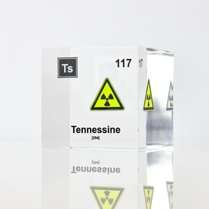Puede incluir: Un cubo de acrílico transparente con el símbolo químico "Ts" y el número atómico "117" impreso en él. El cubo también tiene la palabra "Tennessine" impresa en él, junto con un símbolo de advertencia de radiación amarillo y negro.