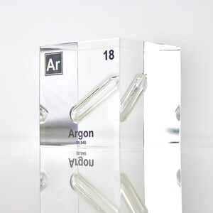 Puede incluir: Un cubo de acrílico transparente con una muestra de gas argón en su interior. El cubo tiene el símbolo químico "Ar" y el número atómico "18" impreso en él. La palabra "Argon" también está impresa en el cubo.