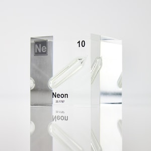 May include: A clear acrylic cube containing a small glass tube filled with neon gas. The cube has the chemical symbol "Ne" and the atomic number "10" printed on it. The word "Neon" and the atomic weight "20.1797" are also printed on the cube.