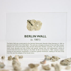 May include: A small piece of the Berlin Wall, a concrete fragment with a rough texture, is displayed in a clear acrylic case. The case also includes a label with the text "BERLIN WALL (c. 1961)" and a brief description of the wall's history.