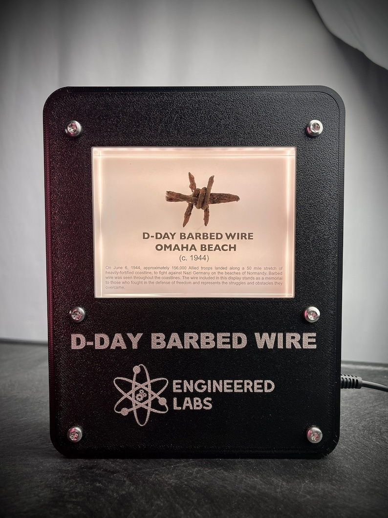 Puede incluir: Una vitrina negra con un frente de vidrio que muestra un trozo de alambre de p&uacute;as de la playa de Omaha durante el D&iacute;a D. La vitrina tiene una placa plateada con el texto "D-Day Barbed Wire" y "Engineered Labs".