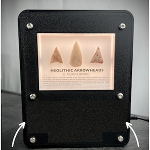 Puede incluir: Una vitrina negra con un frente de vidrio que muestra tres puntas de flecha neol&iacute;ticas. La vitrina tiene un fondo blanco y un borde negro. El texto "NEOLITHIC ARROWHEADS (c. 10,000-3,000 BC)" est&aacute; impreso en una etiqueta blanca dentro de la vitrina. El texto "Customizable Text" est&aacute; impreso debajo de la vitrina.