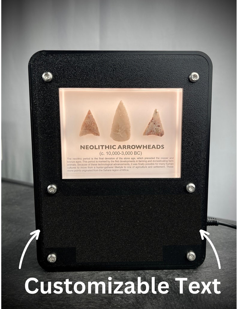 Puede incluir: Una vitrina negra con un frente de vidrio que muestra tres puntas de flecha neol&iacute;ticas. La vitrina tiene un fondo blanco y un borde negro. El texto "NEOLITHIC ARROWHEADS (c. 10,000-3,000 BC)" est&aacute; impreso en una etiqueta blanca dentro de la vitrina. El texto "Customizable Text" est&aacute; impreso debajo de la vitrina.
