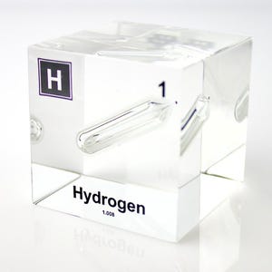 Puede incluir: Un cubo transparente y claro con el símbolo del elemento "H" en un cuadro morado, el número "1" y la palabra "Hydrogen" impresa. El cubo también tiene un tubo transparente similar al vidrio incrustado.
