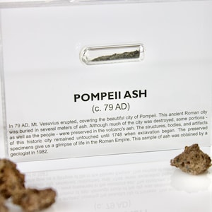 Puede incluir: Un pequeño vial de vidrio que contiene una muestra de ceniza volcánica de la erupción del Vesubio en el año 79 d.C., que sepultó la antigua ciudad romana de Pompeya. La etiqueta dice "Pompeii Ash (c. 79 AD)" y proporciona una breve descripción del evento y su significado.