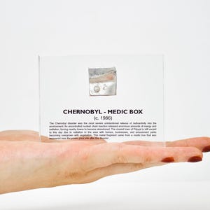 Peut inclure: Une boîte en acrylique transparente contenant une petite boîte en métal argenté. Le texte sur la boîte indique "CHERNOBYL - MEDIC BOX (c. 1986)" et décrit la catastrophe de Tchernobyl. Le texte explique que la catastrophe a été la pire libération accidentelle de radioactivité dans l'environnement. Une réaction en chaîne nucléaire accidentelle a libéré d'énormes quantités d'énergie et de radiations, forçant près de 100 000 personnes à être évacuées. La ville la plus proche de Pripyat est toujours inhabitée à ce jour en raison de la radioactivité dans la zone avec des maisons, des entreprises et des parcs d'attractions envahis par la végétation. Ce petit fragment provient d'une boîte de médicaments qui a été récupérée près de la centrale nucléaire après la catastrophe.