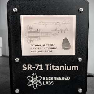 Puede incluir: Una placa de exhibici&oacute;n negra con un marco plateado que muestra un trozo de titanio de un avi&oacute;n SR-71 Blackbird. La placa presenta un diagrama del SR-71 y el texto "TITANIUM FROM SR-71 BLACKBIRD TAIL #61-7970". Debajo del marco, la placa dice "SR-71 Titanium" y "ENGINEERED LABS" con un s&iacute;mbolo de &aacute;tomo.